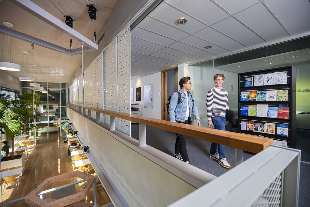 Picture of the inside of TPM faculty building with two guys walking on the mezzanine in front of a rack of journals.